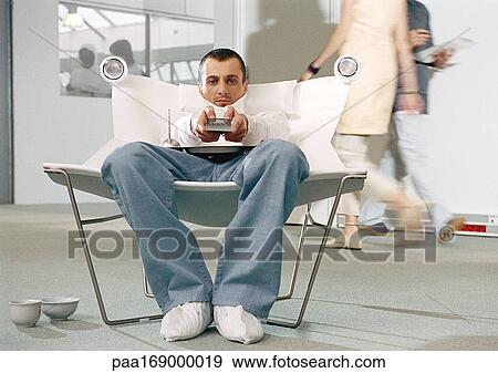 Stock Photo - Man in armchair holding remote control, people in background, portrait. Fotosearch