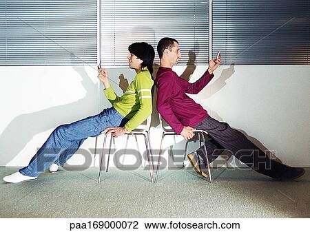 Two men sitting back to back in chairs, using cell phones View Large Photo Image Stock Image - Two men sitting back to back in chairs, using cell phones. Fotosearch