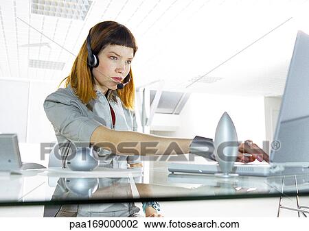 Stock Image - Woman at desk wearing headset in office. Fotosearch