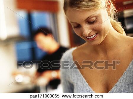 Stock Photography - In kitchen, woman smiling and looking down, man in background. Fotosearch