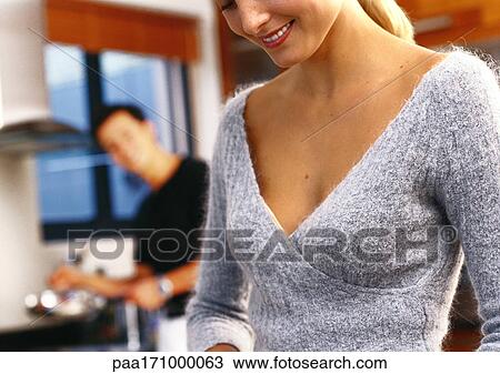 Stock Image - In kitchen, woman smiling, man in blurred background, close-up. Fotosearch