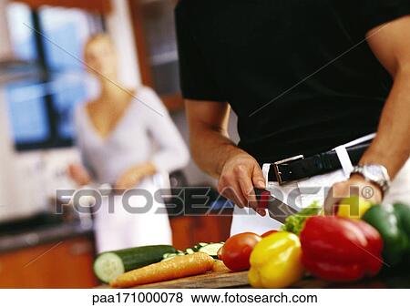 Stock Photo - Man cutting vegetables, mid-section, woman standing in background. Fotosearch