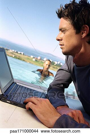 Man using laptop computer, woman in swimming pool in background, tilt View Large Photo Image Picture - Man using laptop computer, woman in swimming pool in background, tilt. Fotosearch