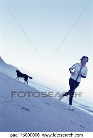 Stock Photograph - Mature woman running on beach, dog in background. Fotosearch