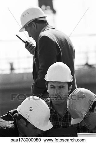 Stock Image - Four men with hard hats, one holding walkie-talkie, b&w. Fotosearch