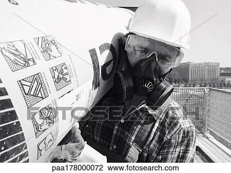 Stock Image - Man wearing hard hat, pollution mask and glasses, carrying roll of insulation on shoulder, close-up, b&w. Fotosearch