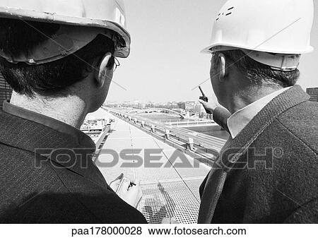 Stock Photo - Two men wearing hard harts, talking, rear view, b&w. Fotosearch