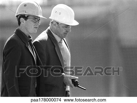 Two men with hard hats, one holding walkie-talkie, waist up, side view, b&w View Large Photo Image Picture - Two men with hard hats, one holding walkie-talkie, waist up, side view, b&w. Fotosearch