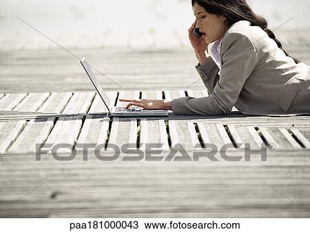 Stock Image - Businesswoman lying on stomach with laptop computer, outdoors. Fotosearch