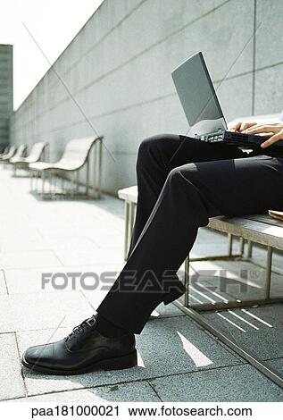 Stock Image - Man on bench with laptop computer on knees, close-up. Fotosearch