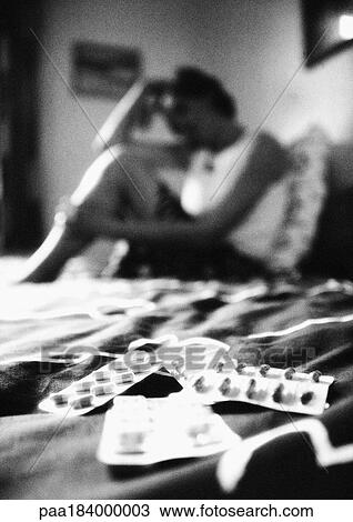 Stock Image - Woman sitting on bed, pills in foreground, blurred, b&w. Fotosearch