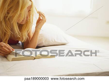 Stock Image - Woman lying on bed reading book, off centered.. Fotosearch