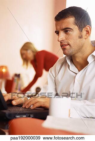 Stock Photo - Man working on computer, woman in background on phone.. Fotosearch