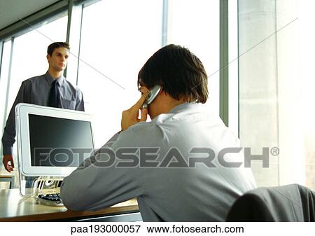 Businessman sitting at computer, using cell phone, second businessman walking in office View Large Photo Image Stock Photo - Businessman sitting at computer, using cell phone, second businessman walking in office. Fotosearch