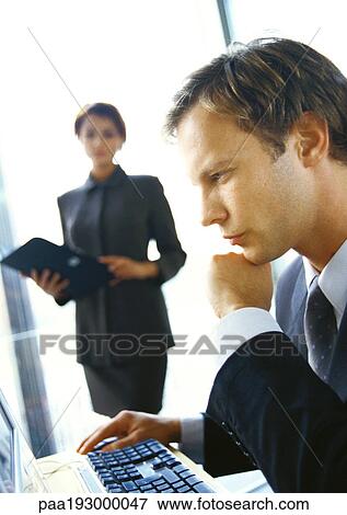 Stock Photo - Businesswoman standing in backgroung, blurred, looking at businessman seated at computer.. Fotosearch