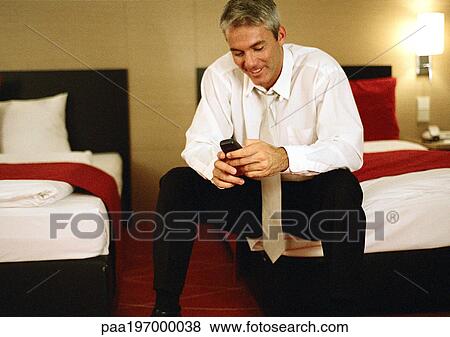Stock Photo - Businessman sitting on bed, using cell phone.. Fotosearch