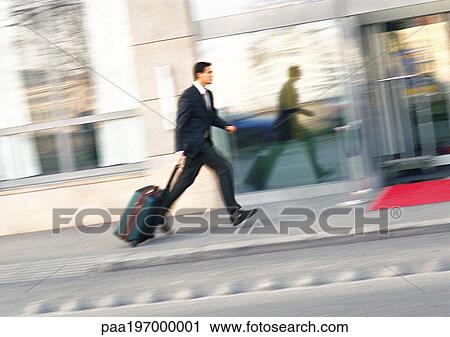 Stock Image - Businessman walking on sidewalk with luggage, blurred.. Fotosearch
