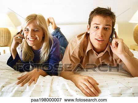 Man and woman lying on their stomachs on bed, using cellphones View Large Photo Image Stock Photo - Man and woman lying on their stomachs on bed, using cellphones. Fotosearch