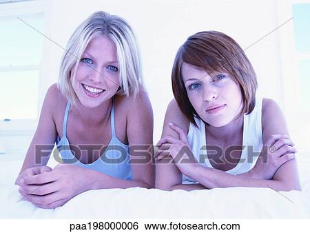 Two women lying on their stomachs on bed, looking at camera View Large Photo Image Stock Photograph - Two women lying on their stomachs on bed, looking at camera. Fotosearch