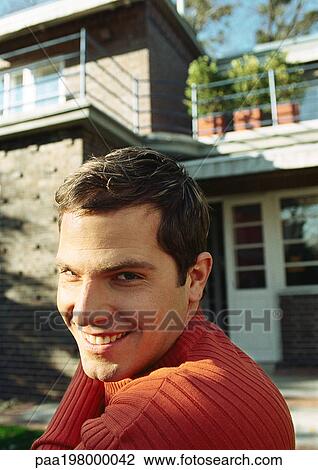 Stock Image - Young man standing in front of a house, smiling at camera. Fotosearch