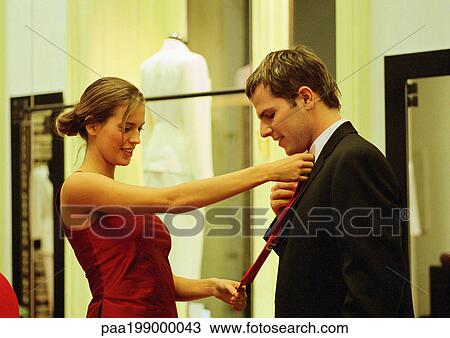 Stock Image - Woman holding neck tie up to man's neck in clothing store, side view.. Fotosearch