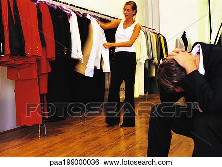 Woman in store looking through clothes, man sitting with head down and hands on neck View Large Photo Image Stock Photograph - Woman in store looking through clothes, man sitting with head down and hands on neck. Fotosearch