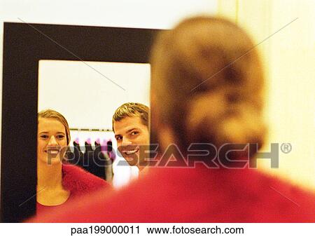 Stock Image - Woman looking in mirror at coat, man's reflection in mirror beside her, view from behind.. Fotosearch