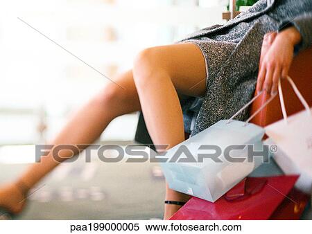 Stock Photography - Woman sitting in chair, leg stretched out, holding shopping bags, low section. Fotosearch