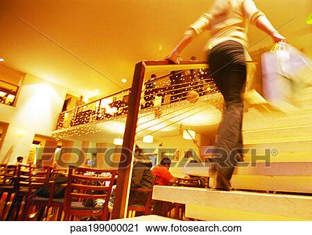 Stock Image - Woman walking up stairs with shopping bags in restaurant setting, blurred.. Fotosearch