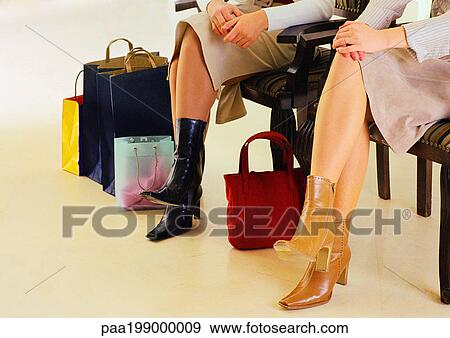 Stock Photo - Women sitting, legs crossed, hands clasped on knees, wearing boots, bags to the side.. Fotosearch