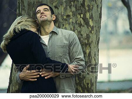 Stock Photo - Man and woman holding each other, leaning against tree.. Fotosearch