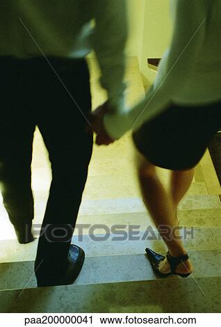 Stock Image - Man and woman walking down stairs holding hands, lower section. Fotosearch