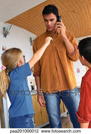 Stock Image - Boy and girl looking up at father, daughter reaching up to father talking on phone.. Fotosearch