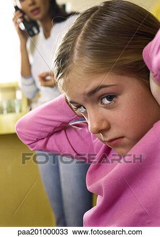 Stock Image - Little girl with hands over ears, looking at camera, close-up, woman standing talking on phone in background. Fotosearch