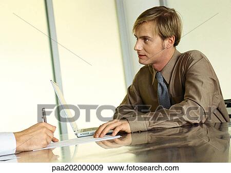 Stock Photo - Businessman sitting at table listening to someone.. Fotosearch