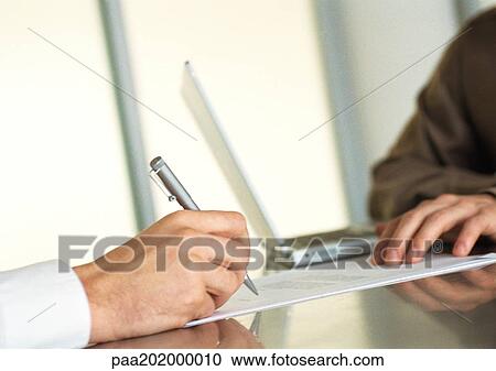 Stock Image - Businessman writing and second business man sitting with laptop, close up of hands. Fotosearch