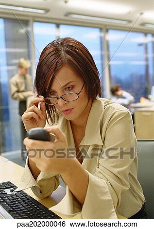 Stock Photograph - Businesswoman adjusting glasses looking at phone, in the office, close up.. Fotosearch