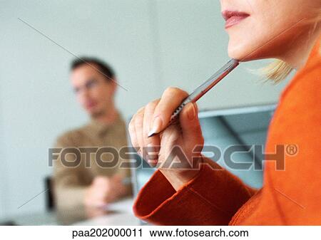 Stock Image - Businesswoman listening holding pen to her chin, man in background. Fotosearch