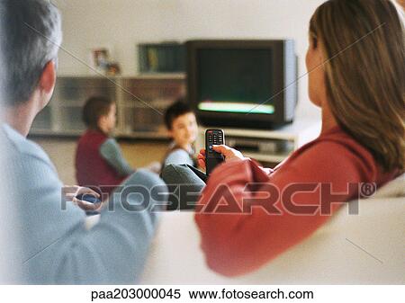 Stock Photography - Adult man and woman sitting on sofa, rear view, blurred in foreground, children near tv in background. Fotosearch