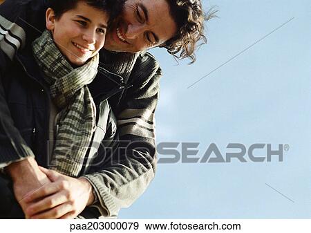Adult man with arms around boy from behind, both in warm clothes, portrait, close up, sky in background View Large Photo Image Stock Photo - Adult man with arms around boy from behind, both in warm clothes, portrait, close up, sky in background. Fotosearch