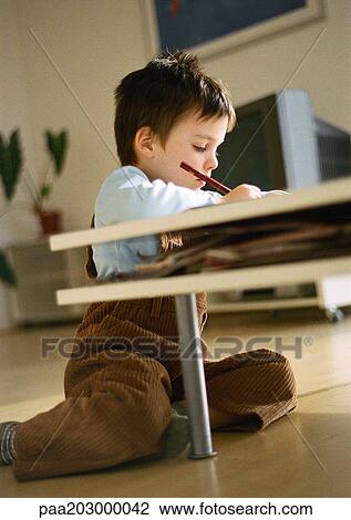 Stock Image - Young boy sitting on floor behind coffee table, drawing, in living room setting, tilt. Fotosearch