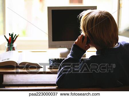 Stock Photo - Young boy sitting at computer talking on phone, rear view. Fotosearch