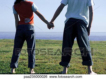 Stock Photograph - Man and woman holding hands on grass by water.. Fotosearch