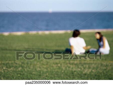 Stock Photograph - Man and woman sitting in grass by ocean, blurred.. Fotosearch