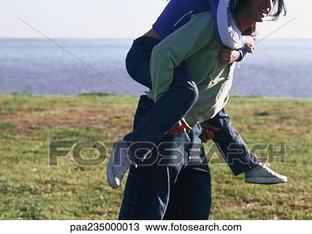 Stock Image - Man carrying woman on his back by the water.. Fotosearch