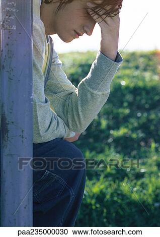Stock Image - Man sitting by pole holding head in front of grass, close up.. Fotosearch