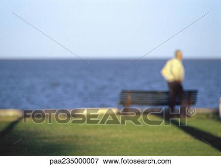 Stock Photo - Man standing in front of bench by the water, blurred.. Fotosearch