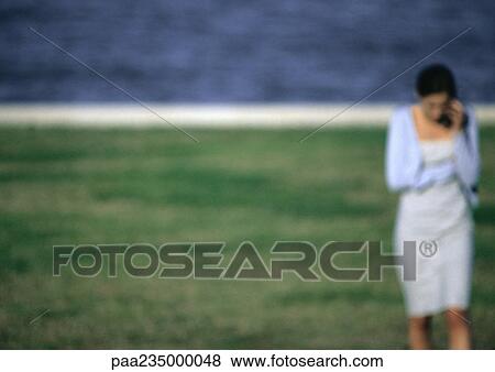 Stock Photo - Woman on cell phone walking on grass by water, blurred.. Fotosearch