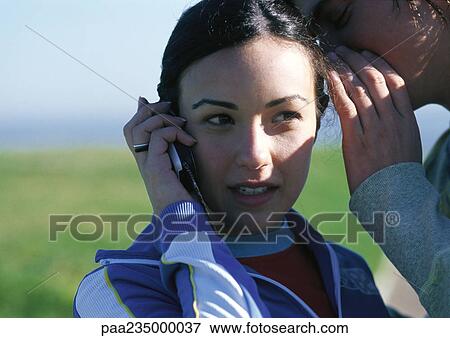 Stock Photo - Woman talking on cell phone with man whispering in her ear, close up.. Fotosearch