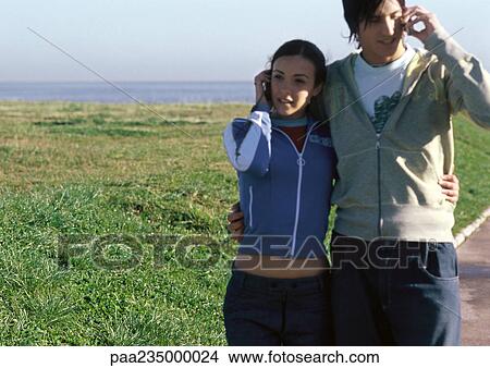 Picture - Young woman and young man talking on cell phones, walking on sidewalk. Fotosearch